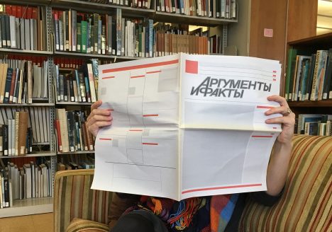 Person holding blank newspaper-style page with title in cyrillic, sitting on couch.