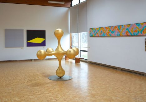 An installation view of Usdan Gallery with work from the "Unpacking the Vault" show installed, including five abstract paintings and a gold colored abstract sculpture