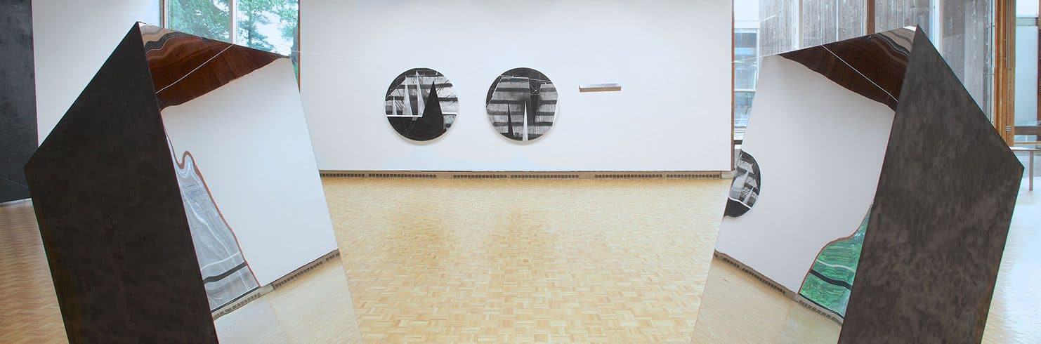 usdan gallery with several works by Torkwase Dyson: two circular canvases and sculptural work of two quadrilateral prisms with one side that is a mirror facing each other.