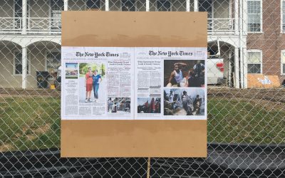 board bolted to chain link fence with new york times article what pasted to it. the newspaper article is marked with highlights and annotations.