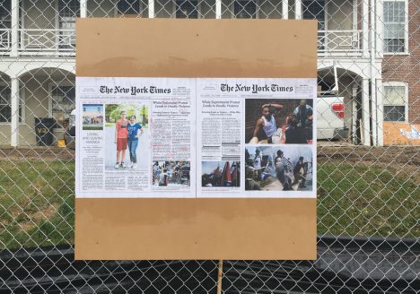 board bolted to chain link fence with new york times article what pasted to it. the newspaper article is marked with highlights and annotations.