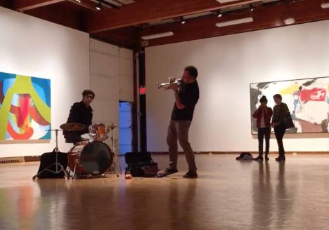 Installation photo of a drummer and a trumpet player. Two viewers watch from the left. In the background, there are two abstract paintings. The one on the right is blue, red, and green. The one of the left is mostly white with red, yellow, and black markings.